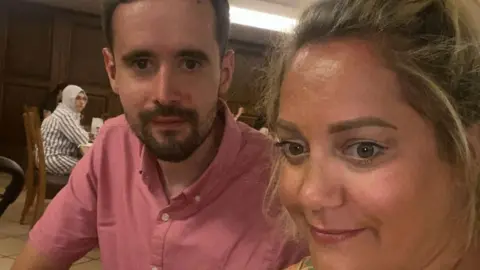 Supplied A man with dark brown hair and a brown beard wears a pink button up shirt, ad a woman with blonde hair both look into the camera.