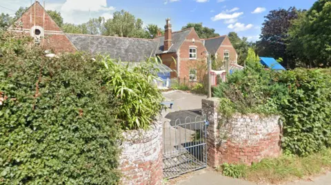 A red brick building, that is East Ruston Infant school and Nursery, is on the Bushes stand next to a silver gate in the middle outside the front of the school. 
