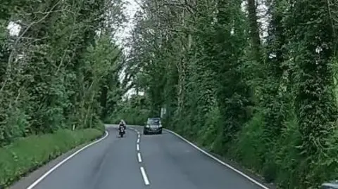 A section of the TT course which has a broken white line running along the middle of it and is lined with green trees. There is a car traveling on the road, with a motorcycle in the other lane travelling in the opposite direction.