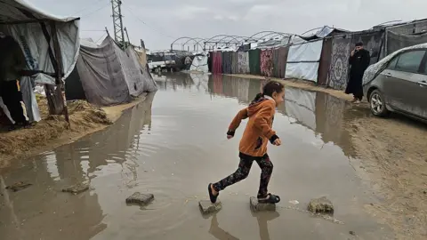 Anadolu via Getty Images A child crosses a flooded track in Khan Younis between makeshift tents used by displaced Palestinians