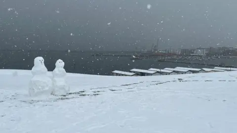 Two snowmen next to each other with a port in the background. Large snowflakes are falling and the ground is covered with thick snow