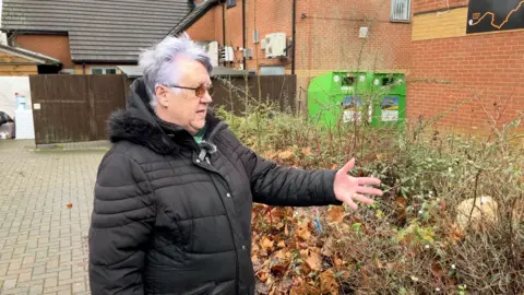 A woman with short purple-white hair, wearing a black winter coat, gestures to an area that is overgrown with scrubby bush and full of litter.