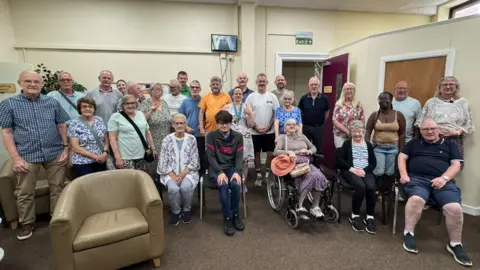 Deaf Connextions A group of around 20 people stand in a room with cream coloured walls looking at the camera. One of them is a wheelchair user. They are a range of ages some young some old. 