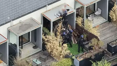 Police officers arresting a man who is on a roof. There are gardens on either side of the one that has several police officers in.