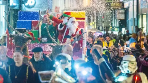 Parish of St Helier Crowds of people surrounding a Christmas sleigh with Santa and an elf on top. Fake snow is being shot out of the sleigh.