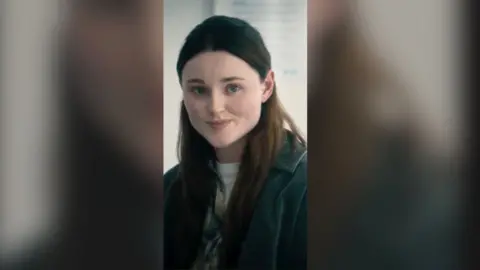 A young woman with long dark hair smiles to someone off screen.