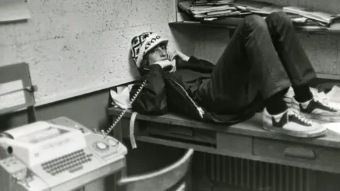 Lakeside School A Bill Gates in teenager wears a hat, lying on the table, tends on the wall with a ground phone, in a black and white image