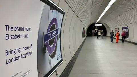 A tunnel in the Elizabeth line system with people walking along in the background. In the foreground is a poster advertising the opening of the new Elizabeth line.