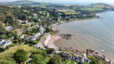 An aerial view of Rockcliffe with water to the right and a bay washing in to a rocky and sandy shoreline. There are houses facing the bay and hills in the distance.
