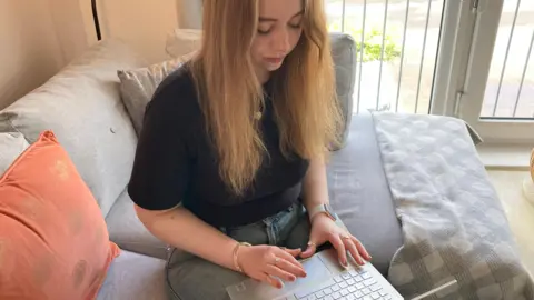 A white woman with long medium blonde hair is typing on her laptop and looking down. She is wearing a black short-sleeved top and jeans, and is sitting on a sofa. 