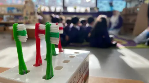 In the foreground there is a toothbrush holder.  It contains three green and two red toothbrushes. In the background, slightly out of focus, are a group of school children who are sitting on a rug and looking towards their teacher.