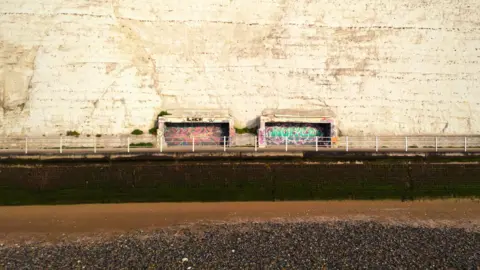 Two concrete shelters sit inside a white cliff. They are derelict and covered in graffiti. 