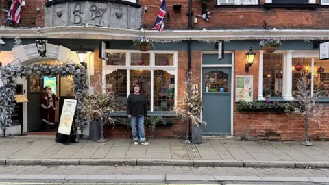 A woman standing on the pavement outside a pub that is covered in Christmas decorations. Santa is in the doorway. She is wearing jeans and a dark jumper.