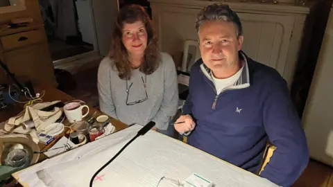 A woman and man sit at a desk with a light overlooking pages of notes on the table. They are sitting in darkness with the light pointing towards the desk.