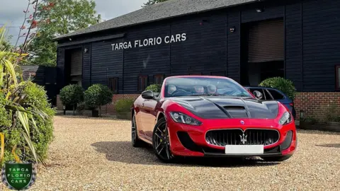 A red open top sports car on gravel in front of a black timber and brick warehouse.