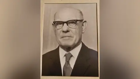 Baroness Golding Ness Edwards, a man with black rimmed glasses, slightly frowning at the camera. He is wearing a black suit with a white shirt and a dark-coloured tie