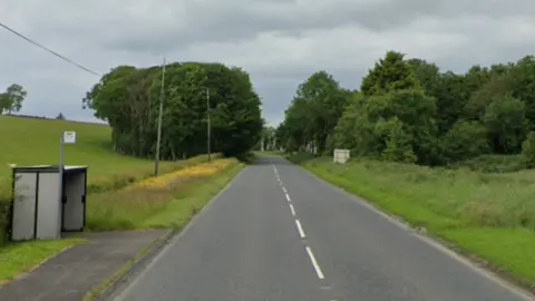 A rural road, to the left there is a bus stop, fields and trees. On the right there are trees and fields 