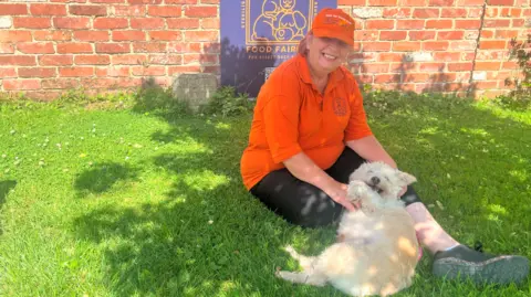 Sammy Cullis sits on the grass with a small white dog and wears an orange Food Fairies-branded polo top and hat. She is in front of a brick wall that bears the charity's logo