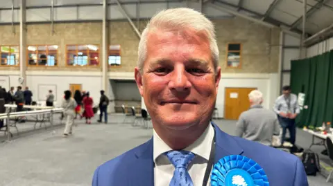 A man in a blue blazer, blue tie, white shirt with a blue rosette stands in a hall looking at the camera. 