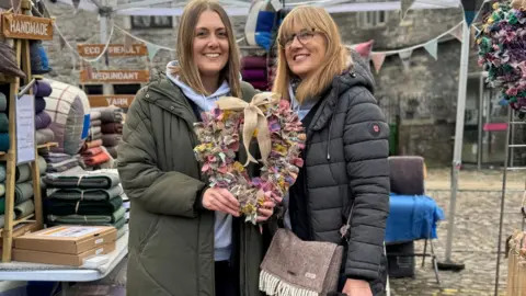 Two women stand in a market place holding a rag wreath in the shape of a heart.