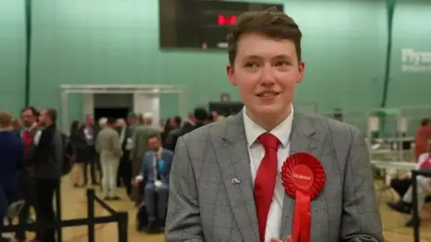 Dylan Tippetts in a white shirt, red tie and grey check suit with a Labour rosette