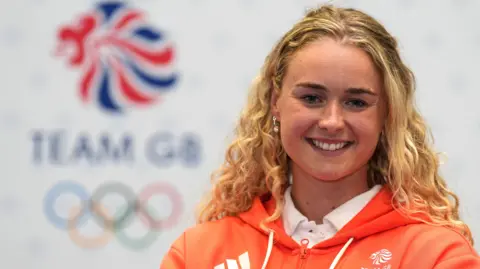 PA Media Hannah Scott smiling at the camera. She is wearing an orange hoodie and a white polo shirt. Behind her is the Team GB logo and the Olympic rings.