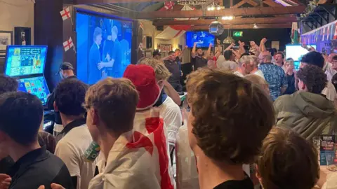 A large crowd of football fans at a pub in Chippenham, Wiltshire. They are all watching the game on a large screen. England flags can be seen dotted around the room.