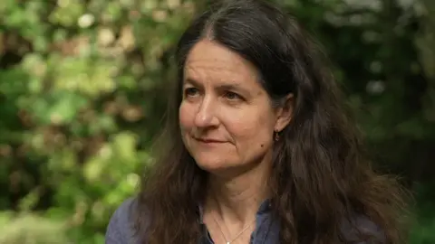 A woman with long dark hair is looking from right to left across the screen. She is wearing a blue shirt and a silver necklace with small earrings. She is sitting in front of a green background of out-of-focus vegetation. 