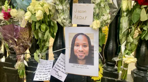 Floral tributes attached to black railings. There is a picture of Jason Arday, with notes written to him. 