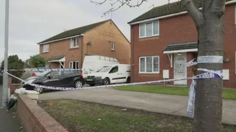 A police cordon in front of a house on Cefndy Road, Rhyl. The tape is tied around a tree and streetlamp in front of the drive. The red brick house has cars parked up to the left of it.