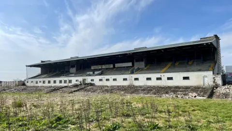A derelict Casement Park, the grass is overgrown and there is rubble in and around the stand