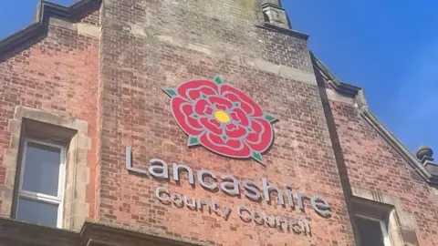 BBC Lancashire County Council sign, with the red rose emblem, on the side of a tall, brick building. The photograph was taken on a sunny day with clear blue skies.