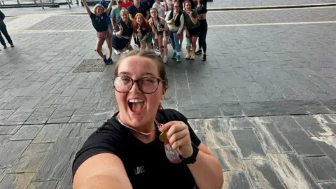 Jade Driscoll Jade taking a selfie portion    posing with a moving  medal. Jade is wearing a achromatic  top, acheronian  rimmed glasses and has an open-mouthed expression. In the photograph  she has besides  included a fig   of chap  runners down  her.