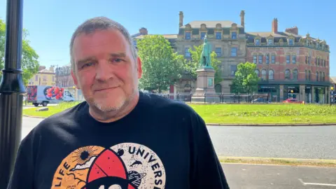 Chris Linton posing for a picture in Barrow. He has short grey hair and blue eyes and is wearing a black t-shirt with a printed logo. He is standing on a pavement with a statue and large building in the background behind him.