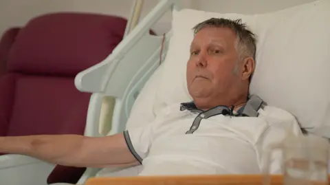 Jabez Petherick, who has short grey hair and is wearing a white polo shirt with grey collar while lying on a hospice bed.