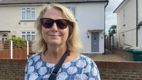 Woman wearing sunglasses smiling at the camera. She is stood outside terraced houses, which are painted cream and white. 