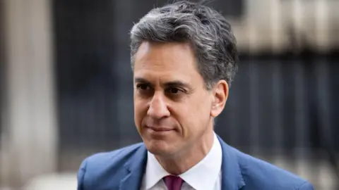 PA Media Ed Milliband standing in Downing Street. He has grey hair and is earing a blue blazer and purple tie.