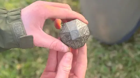 Molly Smith/BBC A 26-sided, intricately engraved grey sword pommel. It is being held in both hands with a blurred background of grass.