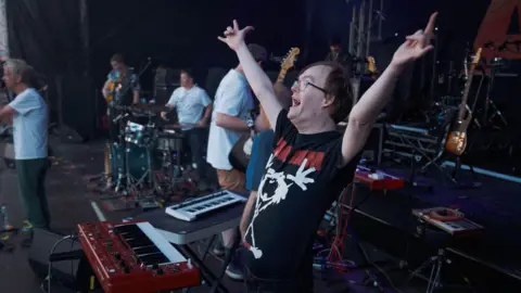 Ben G. Brown A view from the side of the stage, looking at a band set-up. In the background, a group of men can be seen holding a guitar, playing the drums and standing at a microphone. In the foreground, a man stands behind a keyboard and raises his arms in the air, mid-cheer. He has brown hair, and wears glasses and a graphic t-shirt with a white stick figure on it. 