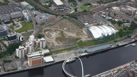 NECA An aerial photo of the Gateshead Quays site which is between the silver, curved Glasshouse building and the brick Baltic building on the edge of the River Tyne. It looks like a building site and is covered in dirt. There are a group of modern apartment blocks behind the Baltic, with office buildings set further back. The white, curved Millennium Bridge goes across the river in front of the arena site.