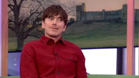 A man, Simon Reeve, has short brown hair and wears a red shirt 