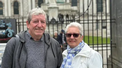A man and a woman standing outside railings, behind them a grand looking white washed Belfast City Hall.
The man has grey hair swept to the side and blue eyes. He is smiling at the camera. He is wearing a grey hoodie over a grey jumper.
The woman is a head or so smaller than him and has white short hair. She is wearing a white coat with a white and blue scarf on. 