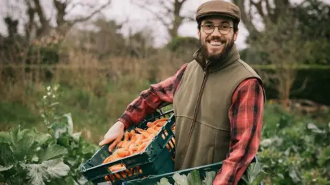 A smallholder wearing a khaki coloured flat cap, gilet fleece and a red checkered shirt beams directly at the camera while stood among green foliage and carrying a box of carrots. 