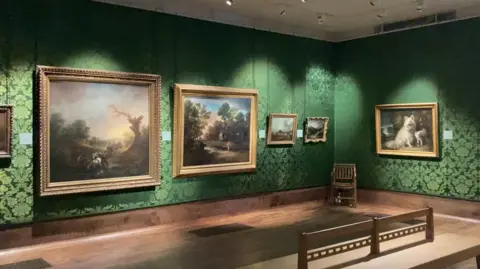 Luke Deal/BBC Large landscape paintings in gold frames in a darkened art gallery, decorated with dark green floral patterned wallpaper 