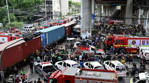 A crowd gathers after a train crashed into a bus at a busy road crossing. There are several red firefighting trucks parked around the train.