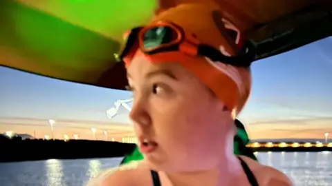 James North A teenage girl looks over her right shoulder while wearing an orange swimming cap and orange goggles as the sunsets behind her. She is stood on a boat as it leaves a port.