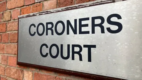 The image shows a silver sign saying 'CORONERS COURT' attached to a red brick wall.