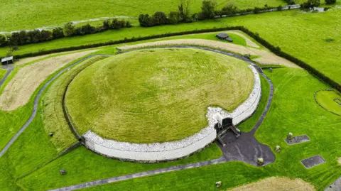 Picture of an ancient burial tomb - it is photographed from above, the tomb is round with a stone wall and grass growing on its roof. It is surrounded by grass and fields. 