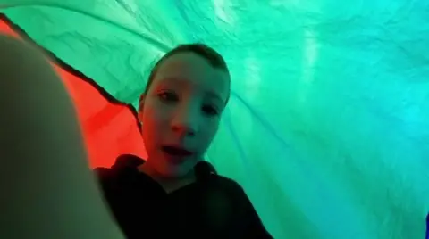 A young boy in a black hoodie with short hair holding a camera in front of him and looking into it. He is underneath a light blue and orange sheet.