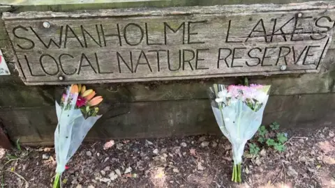 A short of a wooden sign with 'Swanholme lakes Local Nature Reserve carved out' Below are two bunches of flowers.